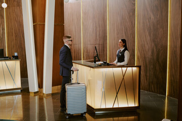 Wide shot of young adult man holding silver suitcase communication with adorable receptionist in...