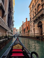 A gondola ride through a narrow Venetian canal, surrounded by historic buildings with ornate facades, reflecting in the calm waters as the boat glides towards a small bridge ahead. © Michael