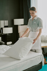 Vertical shot of young adult Caucasian chambermaid in uniform adjusting snow white pillowcase in...