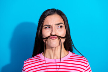 Playful brunette woman wearing striped pink t-shirt creating mustache with hair against blue...
