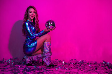Glamorous woman in shiny blue dress holding disco ball against pink background, exuding vibrant...
