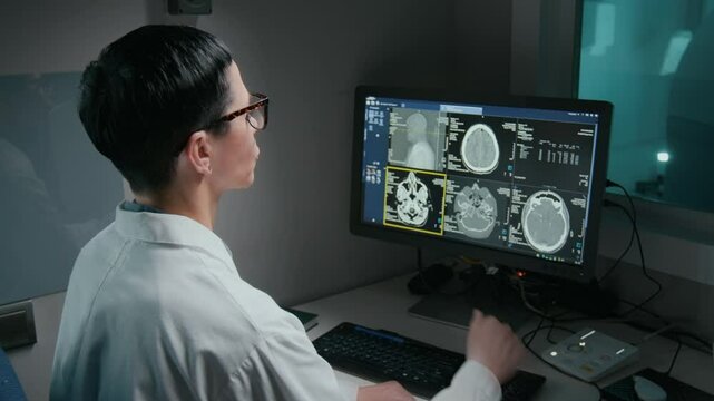 Medium rear shot of female radiographer in white coat, glasses sitting in front of computer, examining brain MRI images, detecting abnormality and scrutinizing it, while at work in hospital