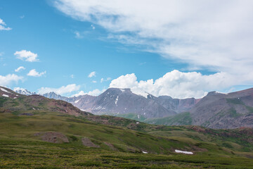 Dramatic alpine view from green hills to large rocky mountain range with glacier under clouds in blue sky. Scenic landscape with big snow mountain top. Changeable cloudy weather in high mountains.