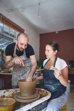 Teaching, man and woman with pottery wheel, clay or lesson for handmade product, learning or talent. Class, creative and skill for production of bowl, instruction or people with equipment in workshop