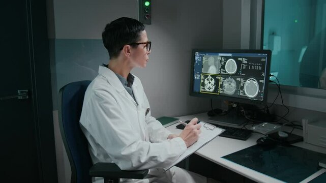 Medium side shot of middle-aged Caucasian female radiography diagnostician in white coat studying patient brain scan on computer screen for abnormalities and writing MRI examination report