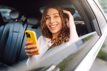 Young woman with curly hair relaxing on the back seat of a car smiling with a phone in her hands. Smiling woman enjoying a taxi ride. Technology and business concept.