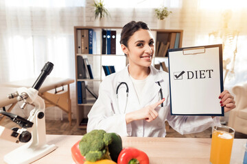 Female doctor sitting at desk in office with microscope and stethoscope. Woman is holding diet sign.