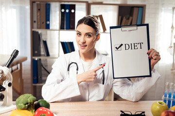 Doctor sitting at desk in office with microscope and stethoscope. Woman is holding diet sign.