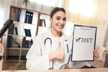 Doctor sitting at desk in office with microscope and stethoscope. Woman is holding diet sign.