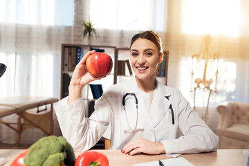 Female doctor sitting at desk in office with microscope and stethoscope. Woman is holding apple and...