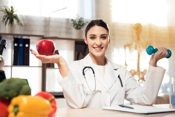 Female doctor sitting at desk in office with microscope and stethoscope. Woman is holding apple and...