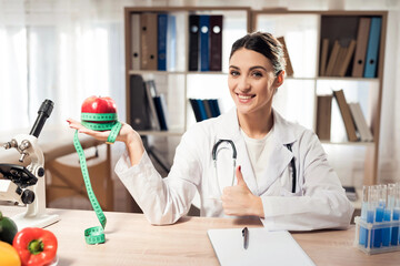 Female doctor sitting at desk in office with microscope and stethoscope. Woman is holding red apple...