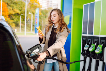A young woman at a gas station fills up her car with one hand and checks her smartphone with the...