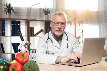 Doctor sitting at desk in office with microscope and stethoscope. Man is working on laptop.