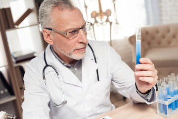 Doctor sitting at desk in office with microscope and stethoscope. Man is holding beaker.