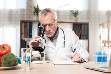 Doctor sitting at desk in office with microscope and stethoscope. Man is looking in microscope.