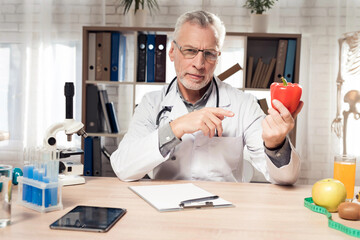 Doctor sitting at desk in office with microscope and stethoscope. Man is holding red pepper.