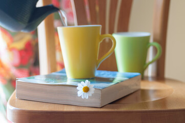 A freshly picked daisy flower used as a book mark in a thick book with hot herbal tea being poured from a cast iron tea pot  into a yellow ceramic mug in the background