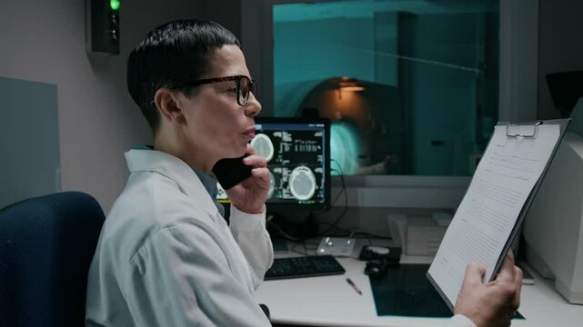 Medium side shot of Caucasian female doctor in white medical coat sitting at desk in clinic, talking to patient on smartphone, discussing positive tomography results, giving treatment recommendations