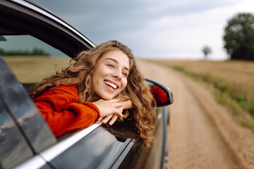 Young woman leaning out of car window, feeling freedom, enjoying warm sunny day. Beautiful woman in...