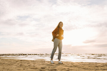 Cute woman with backpack enjoying sunset light on lighthouse, feeling freedom. Tourist relaxing in nature, walking along embankment. Concept of walking, traveling, freedom. Lifestyle.