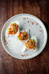 A gourmet dish on a pale plate featuring three crispy shells filled with a colorful mix of diced vegetables, garnished with microgreens and sprinkled with red chili flakes.