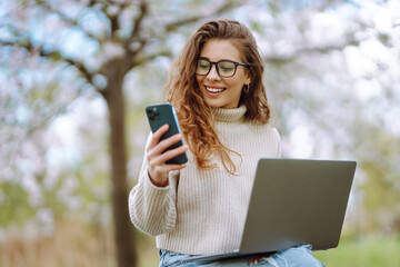 Beautiful woman with curly hair sitting at the table with laptop and phone in blooming park. Young woman working outdoors, using smartphone. Blogging, freelancing concept.