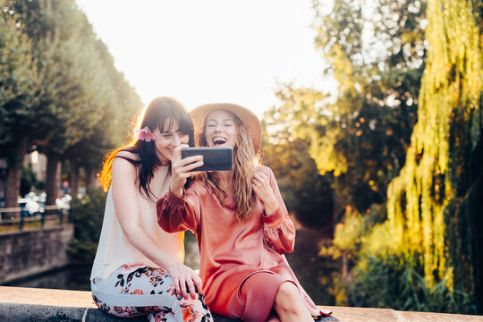 Two happy young women taking a selfie outdoors on a sunny day - Cheerful young women taking a selfie with a smartphone while enjoying a sunny day - Powered by Adobe