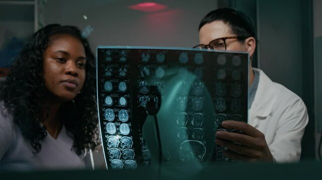 Medium shot of mature Caucasian female radiographer in white coat consulting young black woman in clinic, demonstrating brain PET scans, pointing to abnormalities. MRI machine reflection on glass