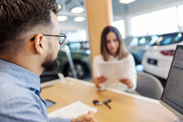 Rear view of car seller sitting at car showroom with customer reading a contract.