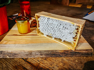 A wooden honeycomb frame filled with fresh, raw honey sits on a rustic wooden table alongside a jar of golden honey. The scene highlights the purity and richness of natural beekeeping.