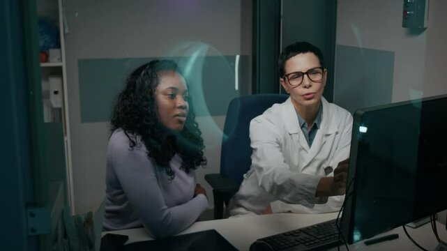 Medium shot, from behind glass window with MRI machine reflection, of cheerful Caucasian female radiology doctor and African American patient examining scans on computer screen, discussing treatment