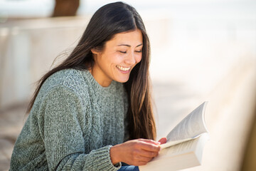 Smiling woman with long hair and cozy knitted sweater reading a book outdoors in natural light