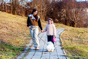 Fototapeta premium Two cute little girls were caught walking with a very fluffy white dog in a green park. The girls were walking with the dogs with enormous pleasure.