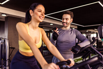 Naklejka premium Woman rides a bicycle while her personal trainer is encouraging her, in the gym.