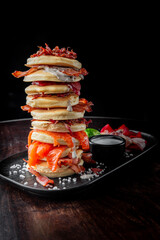Stacked pancakes layered with crispy bacon and salmon, garnished with a creamy sauce. The dish is presented on a black plate against a dark background, creating a striking visual contrast.