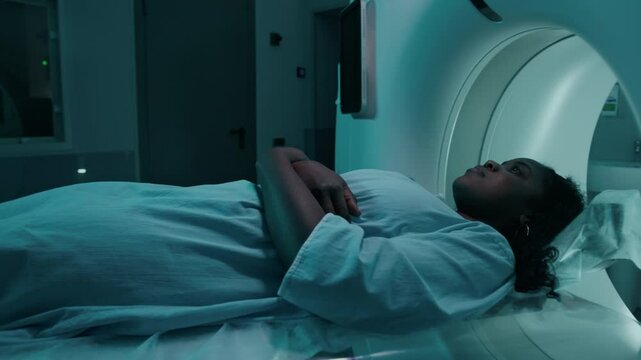 Medium full tracking shot of young African American female patient in disposable hospital gown sliding on table into MRI machine for brain scanning, while diagnostician is watching behind glass window