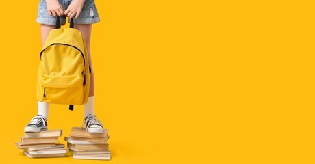 Teenage girl in sneakers and with backpack standing on books against yellow background with space...