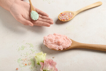 Female hand with wooden spoons of body scrubs and flowers on light background, closeup
