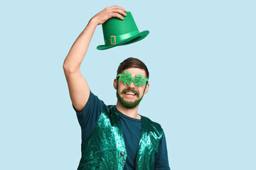 Young man in decorative glasses in shape of clover with green beard holding leprechaun hat on blue...