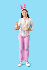 Beautiful young woman in bunny ears holding wicker baskets with Easter eggs on blue background