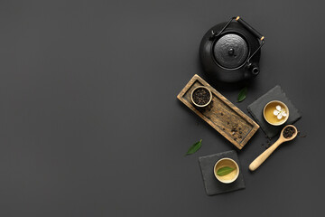Teapot with cups, wooden spoon of dry tea and leaves on black background