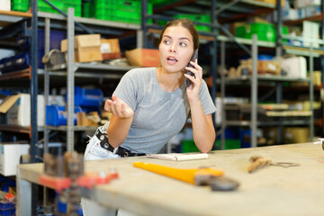 Portrait of young saleswoman confirming order from customer at workplace in warehouse, talking smartphone