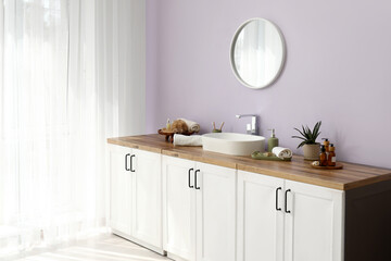 Mirror, chest of drawers with sink and bath supplies near lilac wall in interior of bathroom