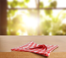 Red checkered tablecloth on wooden table in room against blurred sunlit window, closeup