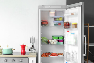 Open fridge full of fresh food near white wall in kitchen