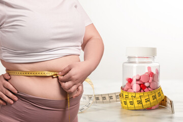 Weight loss. Overweight woman measuring her waist with tape and pills on white background, closeup