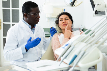 Fototapeta premium Asian woman talking with african-american man dentist about toothache during visit in dental clinic.