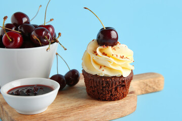 Wooden board with sweet cherry cupcake and bowl of jam on blue background