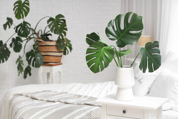 Interior of light bedroom with cozy bed and monstera leaves on bedside table